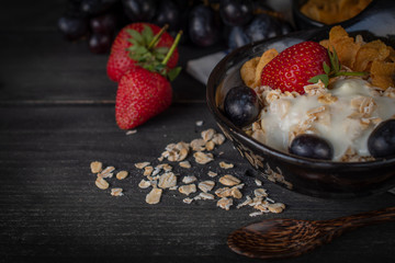 Yoghurt mix oatmeal, strawberry and grape topping in black bowl on wood table with spoon, grapes, strawberry and oatmeal placed around.