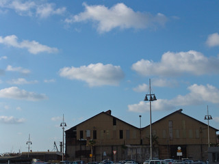 Urban view of two roof tops of big buildings by the sea port, boat houses with tall city lamps in front, under cloudy sky.