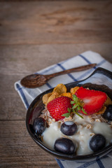 Yoghurt mix oatmeal, strawberry and grape topping in black bowl on Blue and white striped fabric and wood table with spoon placed back side.