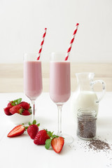 Strawberry smoothie. 2 glasses of strawberry milkshake, glass pitcher of milk, small jar of chia seeds, porcelain bowl with strawberries embellished with few real strawberries and fresh mint.
