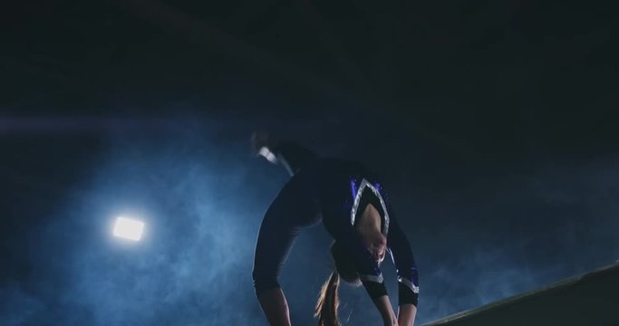 The girl is a professional athlete performs gymnastic acrobatic trick on a beam in backlight and slow motion in sports gymnastic clothing. Smoke and blue. Jump and spin on the balance beam