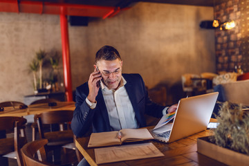 Hardworking middle aged businessman dressed smart casual talking on the phone and looking at documentation while sitting in cafeteria at evening. On a desk in front of him laptop.