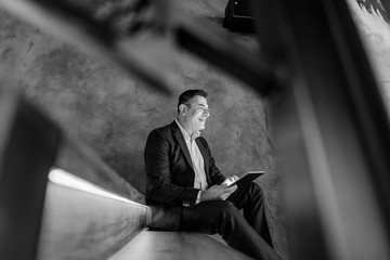 Middle aged businessman sitting on the stairs at cafeteria and using tablet. Black and white photo.