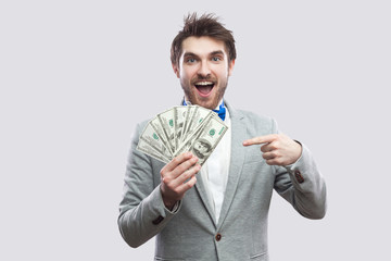 Happy amazed handsome young businessman in classic gray coat and blue bow tie holding many dollars, pointing finger and looking at camera with open mouse. Indoor studio shot, isolated, gray background