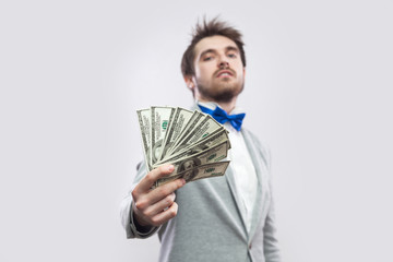 Take it! Portrait of confident attractive young businessman in gray coat and blue bow tie standing, proudly with head up and giving you many dollars. Indoor, studio shot, isolated, gray background.