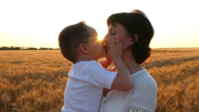 A happy mother kisses a child holding him in her arms in a wheat field on a sunset background