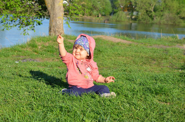 Smiling cute playful little girl is standing on green grass. girl toddler walks around the lake learns to walk. sunny sping or summer day