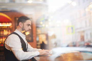 Side view portrait of handsome businessman using laptop in cafe shot from behind glass, copy space