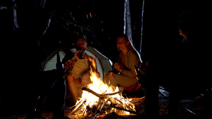 Group of friends drinking beer near bonfire, tent camp, friendly atmosphere