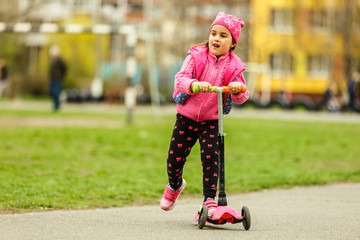 Little child learning to ride a scooter in a city park on sunny summer day. Cute preschooler girl riding a roller. Kids play outdoors. Active leisure and outdoor sport for children.