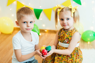 blond, blue-eyed cute smiling children,boy in a white t-shirt and ponytail hair girl in yellow dress, brother and sister 4-5-6 years with Easter eggs in a room on the background of yellow decoration