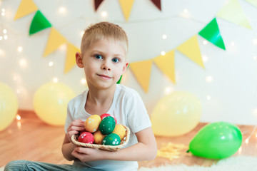 Fototapeta na wymiar blond, blue-eyed cute smiling boy in a white shirt, the age of 6 years, with easter eggs in a room on the background of yellow design, sitting, studio