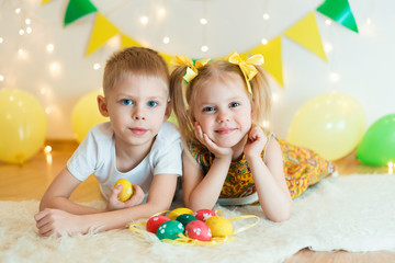 Fototapeta na wymiar blond, blue-eyed cute smiling children,boy in a white t-shirt and ponytail hair girl in yellow dress, brother and sister 4-5-6 years with Easter eggs in a room on the background of yellow decoration