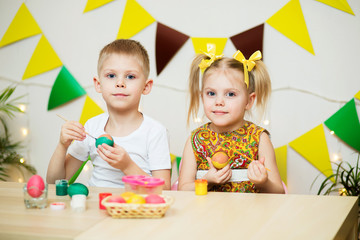 blond, blue-eyed cute smiling children, a boy in a white t-shirt and ponytail hair girl in a yellow dress, brother and sister 4-5-6 years painted Easter eggs in a room