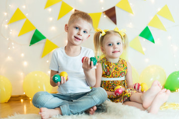 blond, blue-eyed cute smiling children,boy in a white t-shirt and ponytail hair girl in yellow dress, brother and sister 4-5-6 years with Easter eggs in a room on the background of yellow decoration