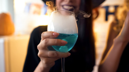 Young lady smiling and holding blue cocktail with white smoke at Halloween party