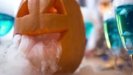 Carved jack lantern smoking, blue cocktails on Halloween party table, close-up