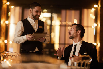 Portrait of mature gentleman ordering food in luxury restaurant with smiling waiter looking at him,...