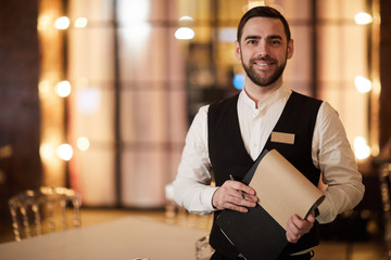 Waist up portrait of handsome waiter smiling cheerfully at camera standing in luxury restaurant or cafe, copy space - obrazy, fototapety, plakaty