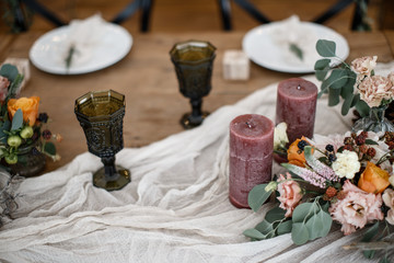 Couple of wineglasses and a couple of a candles are standing on the table alongside with some flowers in a pots.