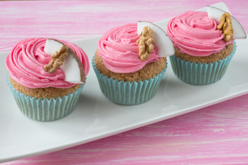 Three pink cupcakes decorated with whipped cream, piece of coconut and piece of walnut on a white ceramic plate standing on a wooden pink background