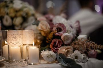 A lot of flowers are laying on the table alongside with some candles in a candle glasses.