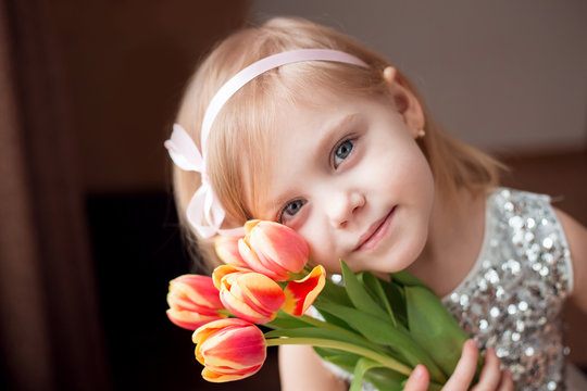 Little blonde girl with a bouquet of tulips., pressed to the colors in a white dress with a ribbon in her hair, horizontal image, look at the camera, with a gentle smile