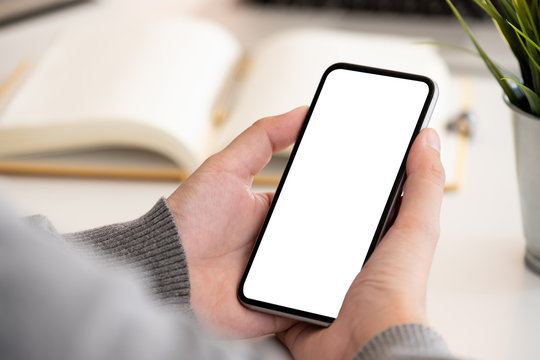 Mockup image of hand holding black mobile phone with blank white screen on workplace desk.