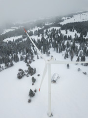 Aerial view of wind turbine in snow covered landscape in Swizterland. Tall pylon in fog, fir trees in the background.