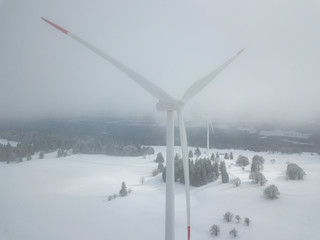 Naklejka na ściany i meble Aerial view of wind turbine in snow covered landscape in Swizterland. Tall pylon in fog, fir trees in the background.