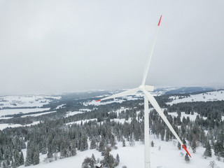 Aerial view of wind turbine in snow covered landscape in Swizterland. Tall pylon in fog, fir trees in the background.