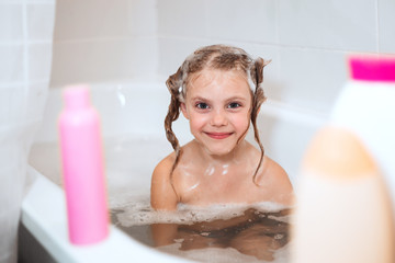 happy  little girl of 6 years old (babe) sits in the bathroom in the water with fluffy foam, washes her head herself using a shampoo without tears that does not pinch the eyes