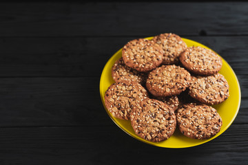 Oatmeal cookies with seeds and sesame seeds on a yellow plate, on a black wooden background