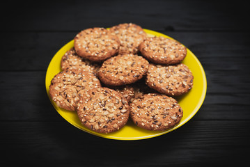 Oatmeal cookies with seeds and sesame seeds on a yellow plate, on a black wooden background