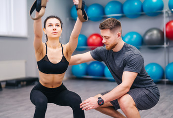 Fototapeta premium Muscular beardy trainer helping attractive female to practice with TRX in the fitness center. Sporty woman doing exercises with personal instructor at gym. Close-up