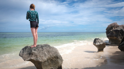 Teenage girl standing on single rock observing the beach, framed left, looking right
