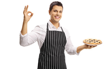 Smiling young man with an apron holding a pie and making a sign symbolizing