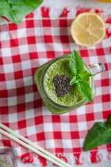 Top view Healthy green spinach smoothie in a jar mug decorated with mint and chia seeds with ingridients on the checkered napkin on the white marble table. Selective focus.