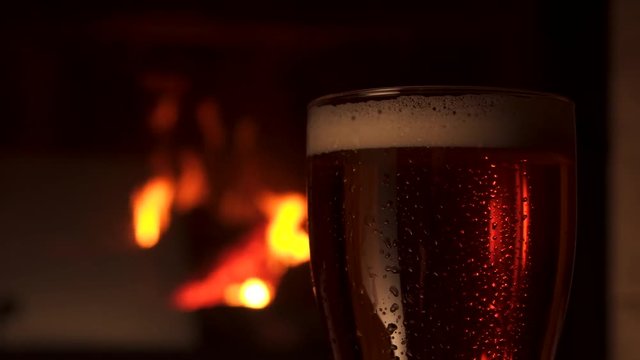 Close-up transparent glass bocal with water drops of refreshment cooled beer with foam fireplace in background. Big mug of fresh craft alcohol beverage at evening cosiness atmosphere