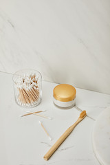 ear sticks in glass, cosmetic cream and bamboo toothbrush on white marble background