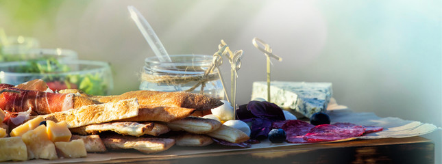 Tray with bacon, cheese cubes, salami, ham; decorated catering table. Nature spring or summer background