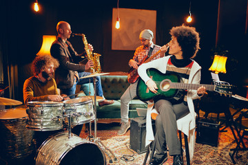Mixed race woman playing acoustic guitar and sitting with legs crossed in home studio. In background the rest of the band playing drums, saxophone and bass guitar.