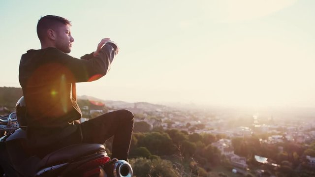 Traveler and cool biker man sitting on moto and creating content for his social network like Instagram or facebook taking picture or photo of sunrise or sunset cityscape flare by smartphone or mobile