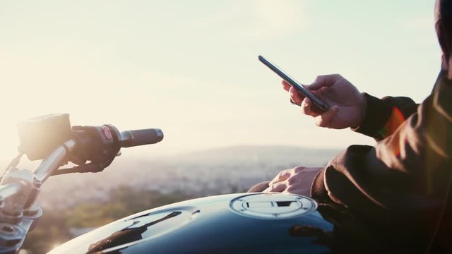 Motobiker man using mobile phone or smartphone and cheking or chatting or texting or googling on social network like instagram or facebook feed on sunrice or sunset outdoors conected to internet 4g