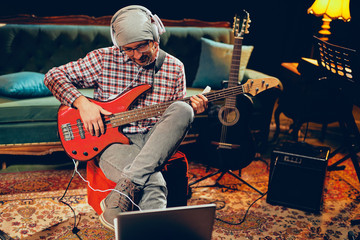 Young Caucasian man with hat on head playing bass guitar while sitting in home studio.