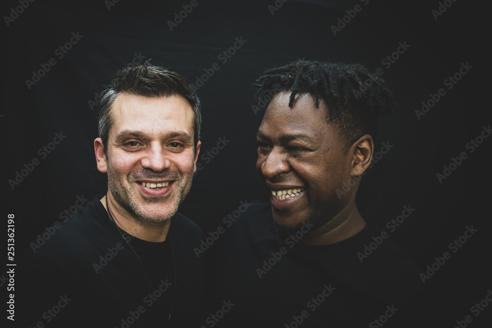 Wall mural An interracial gay couple pose before the camera to have their pictures taken - black man and mixed race man with stubble - on a black background - with copy space.