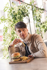handsome young man holding french fry near tasty burger on cutting board in cafe