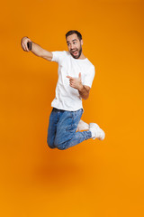 Fototapeta na wymiar Full length photo of brunette guy 30s in casual wear laughing and taking selfie on cell phone, isolated over yellow background
