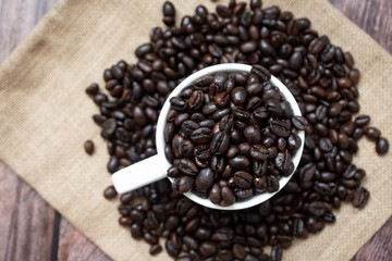 Many coffee beans inside cup of coffee and on sackcloth placed on wooden table. Top view focus.
