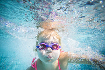 Wide angle underwater photo of a toddler girl swimming in a big swimming pool with goggles and a...
